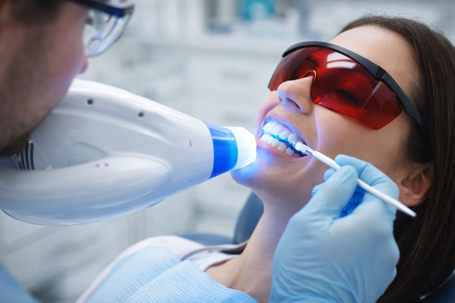 Laser Teeth Whitening London treatment in progress, close up of patient wearing protective eyewear while dentist applies whitening gel with blue activation light in modern clinic.